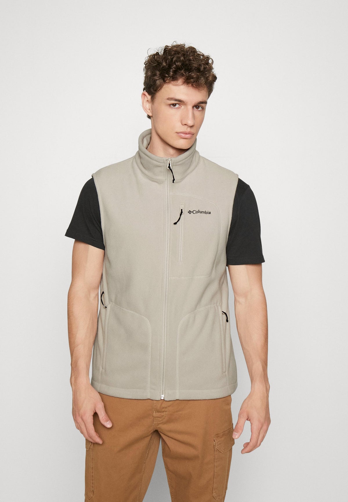 A man wearing a beige Columbia Men's Fast Trek Vest with a black T-shirt underneath and brown pants.