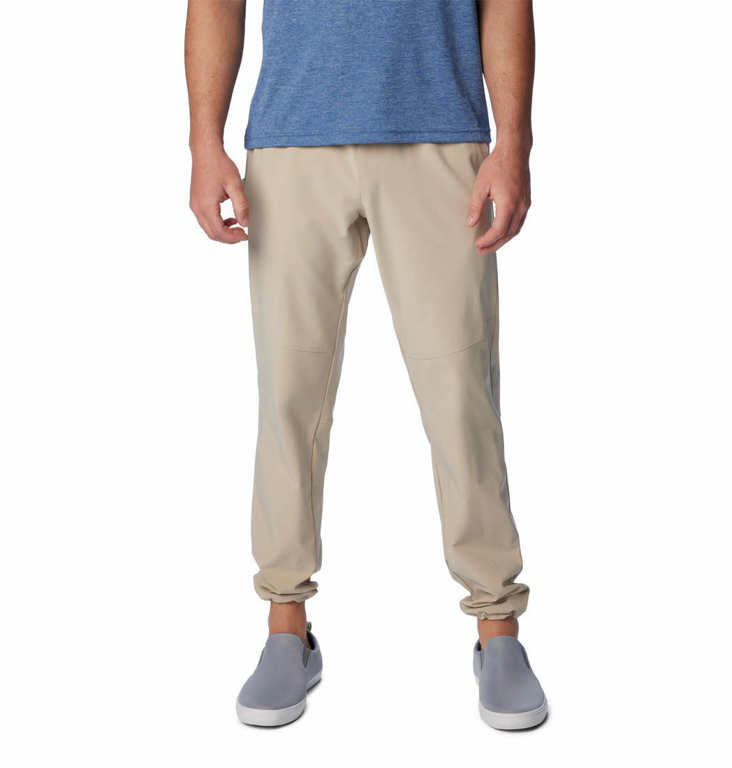 A man standing against a white background, wearing beige Columbia men's Omni-Shade Terminal Roamer Stretch Pant and a blue t-shirt with gray sneakers.