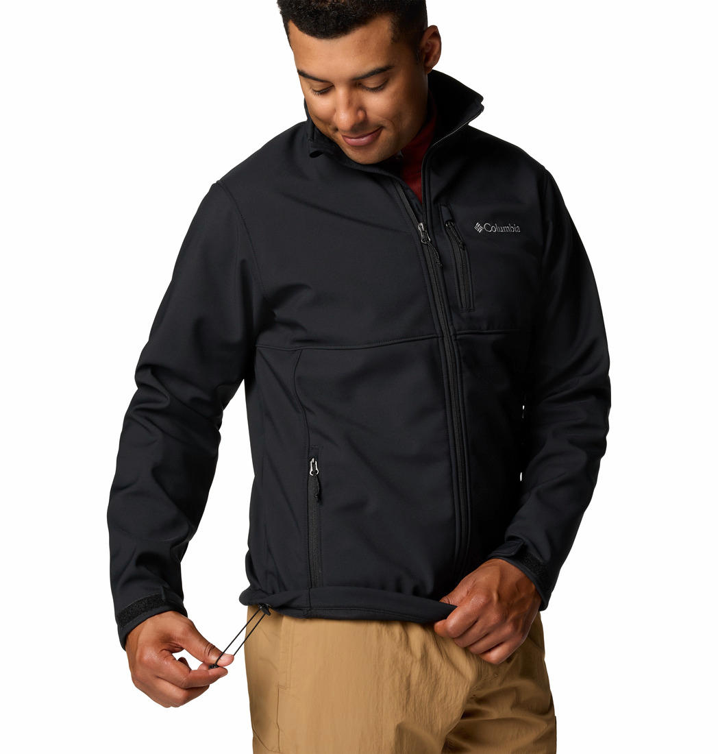 A man wearing a black Columbia Men's Omni-Shield Ascender Softshell Jacket with a zip-up front, hand pockets, and a drawcord adjustable hem.