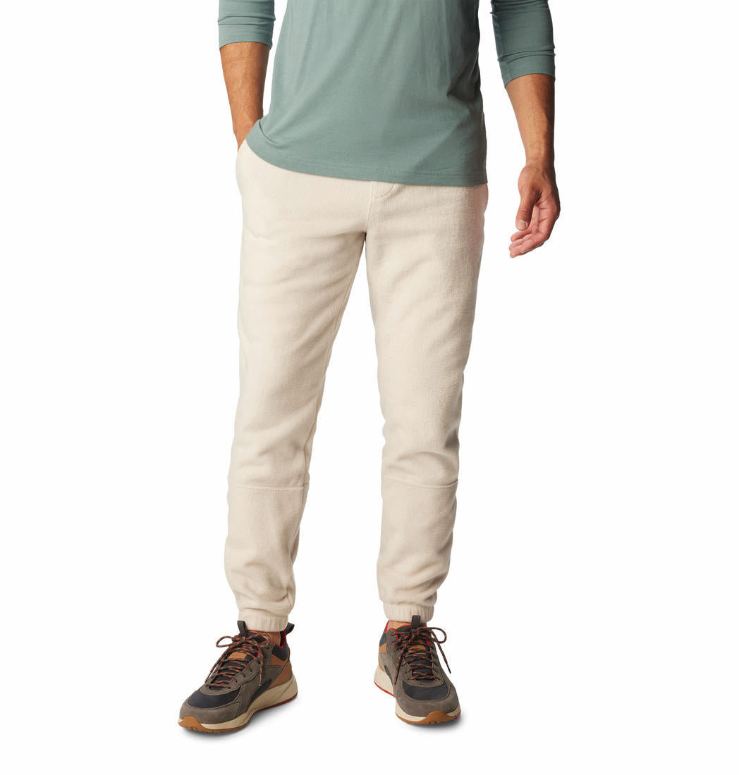 A man standing with hands in pockets wearing beige fleece pants and a green shirt with brown shoes.