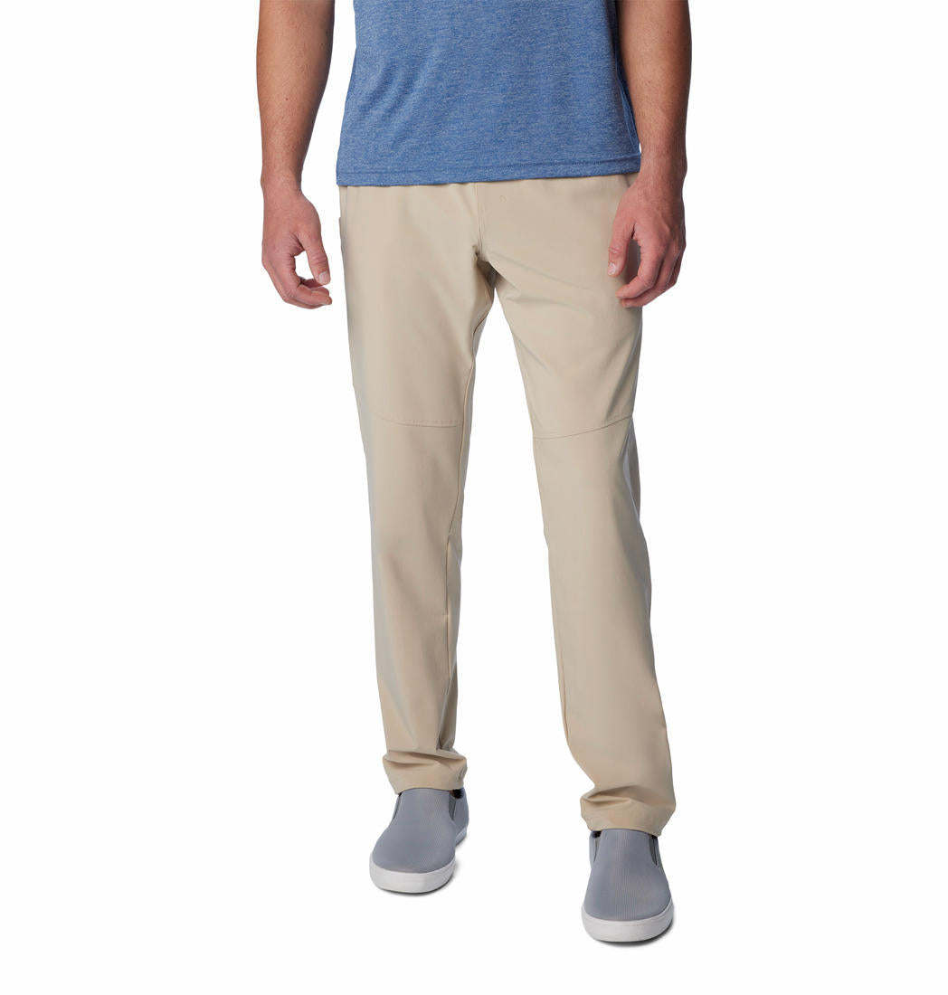 A man standing against a white background, wearing beige Columbia men's Omni-Shade Terminal Roamer Stretch Pant and a blue t-shirt with gray sneakers.