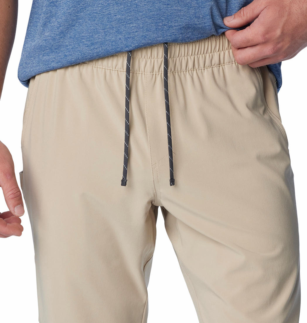 A man standing against a white background, wearing beige Columbia men's Omni-Shade Terminal Roamer Stretch Pant and a blue t-shirt with gray sneakers.