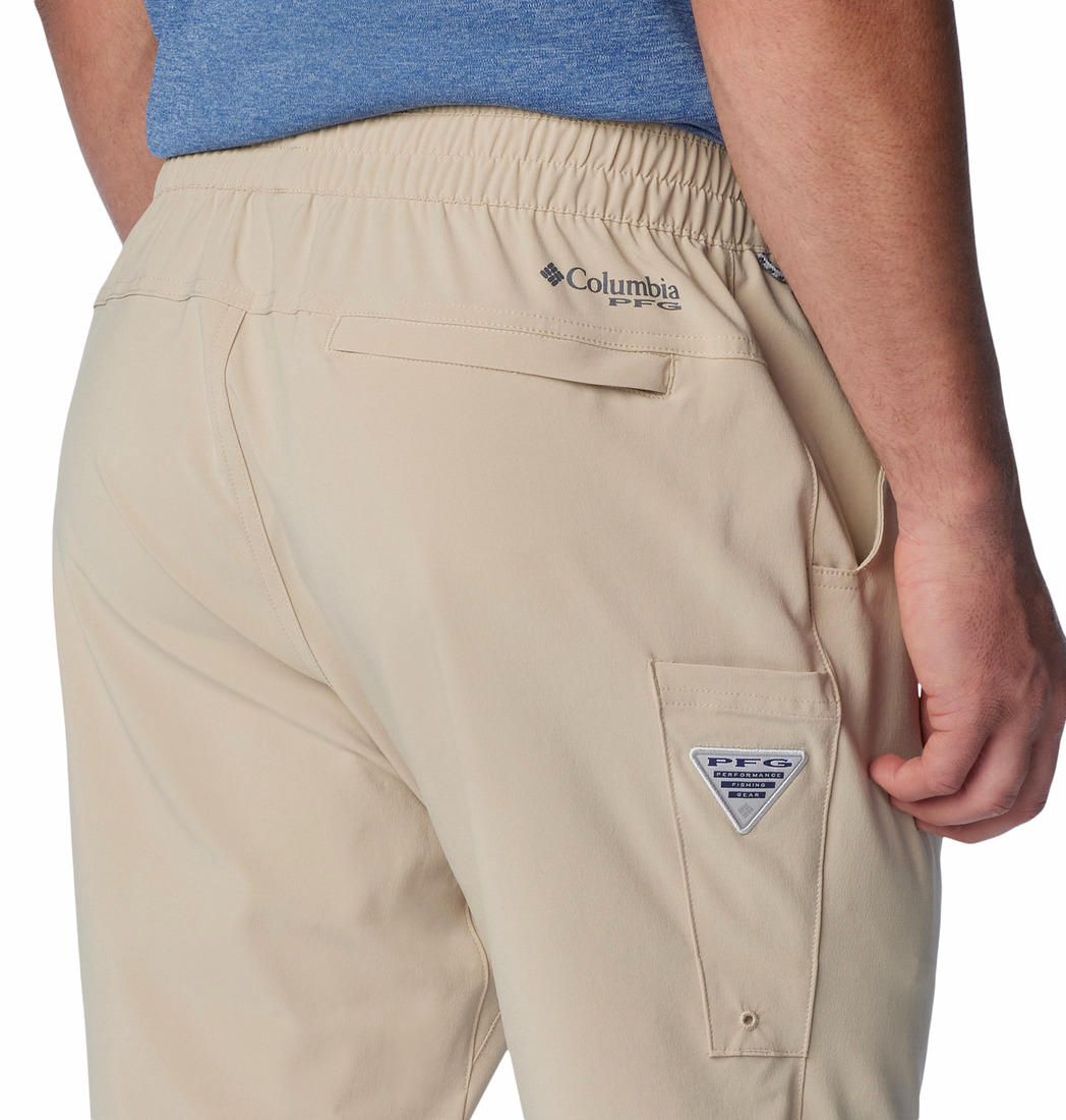 A man standing against a white background, wearing beige Columbia men's Omni-Shade Terminal Roamer Stretch Pant and a blue t-shirt with gray sneakers.