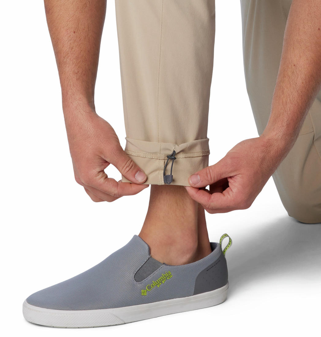 A man standing against a white background, wearing beige Columbia men's Omni-Shade Terminal Roamer Stretch Pant and a blue t-shirt with gray sneakers.